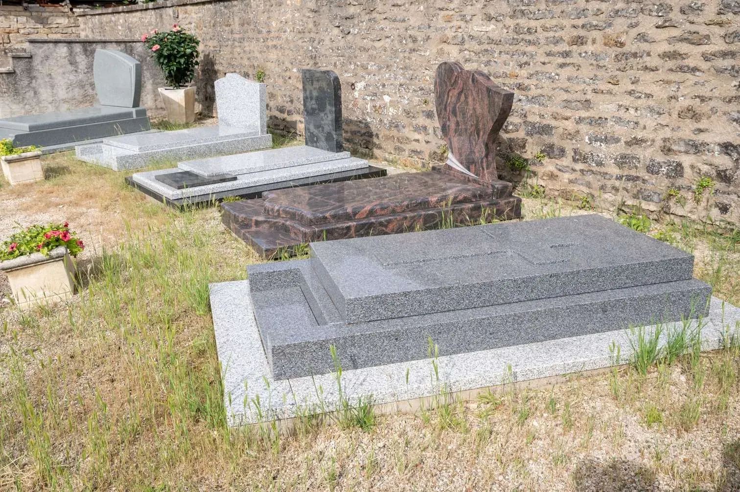 Exposition en plein air de tombes et monuments funéraires en granit le long d'un mur en pierre.